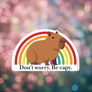 May include: A sticker featuring a brown capybara with a rainbow arch. The text "Don't worry. Be capy." is below the capybara. The background is a blurred mix of pink and green.