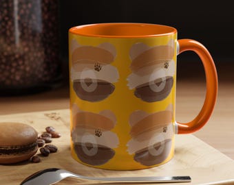 Puede incluir: Taza de cerámica amarilla con asa e interior naranja. La taza está decorada con un patrón repetido de diseños en forma de oso en tonos marrones y beige. Una huella de pata es visible en cada diseño de oso.