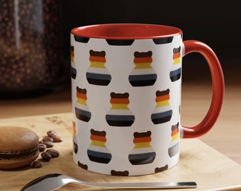 Puede incluir: Una taza de cerámica blanca con interior y asa rojos. La taza está decorada con un patrón repetido de diseños estilizados en forma de oso de colores. Una cuchara y un pastelito están en primer plano, descansando sobre una superficie de madera.