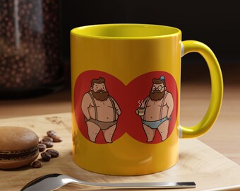 Puede incluir: Taza de cerámica amarilla con asa e interior amarillos. La taza presenta un diseño rojo en forma de corazón con dos hombres de dibujos animados en ropa interior. Un hombre sostiene una taza de café. Una cuchara y un macaron están en primer plano.