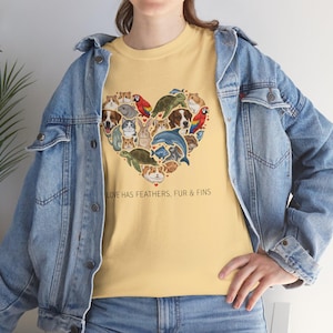 May include: A light yellow t-shirt featuring a heart-shaped design composed of various animals, including dogs, cats, birds, and a dolphin. The text "LOVE HAS FEATHERS, FUR & FINS" is printed below the heart. The person is wearing a blue denim jacket.