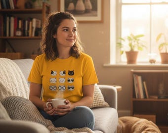 May include: A woman wearing a yellow t-shirt featuring a grid of cat faces with various expressions. She is seated on a grey sofa, holding a white mug. A dog is lying on the floor near a window with potted plants.