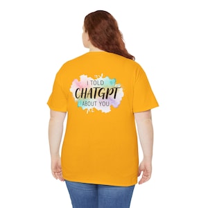 May include: A mustard yellow short-sleeve t-shirt featuring a watercolour-style graphic on the back. The design includes a pastel-coloured splash background with the black text "I TOLD CHATGPT ABOUT YOU" printed in the centre of the graphic.