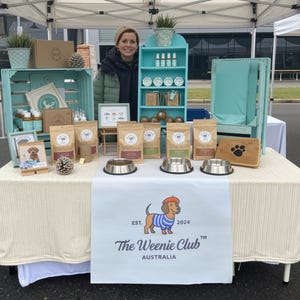 May include: A display table at an outdoor market features dog food, bowls, and accessories. The table is decorated with a banner that reads "The Weenie Club AUSTRALIA." A woman stands behind the table, smiling. The products are arranged on a white tablecloth.
