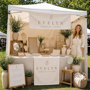 May include: A jewelry studio booth featuring a white canopy, a table draped in beige fabric, and a banner reading EVELYN JEWELRY STUDIO. The display includes various jewelry stands, necklaces, earrings, and greenery in vases and baskets.