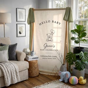 May include: A beige baby shower banner with the text "HELLO BABY" and "Grace's BABY CELEBRATION" with a teddy bear illustration. The banner is supported by a wooden frame and decorated with green fabric.