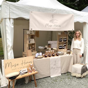 Peut inclure: Un stand de marché présentant les produits "Muse Homes Soy Candles". Le stand comprend une bannière, des caisses en bois et une table avec des bougies, des savons et d'autres articles. Une femme se tient près de la table, et un petit banc en bois expose d'autres produits.