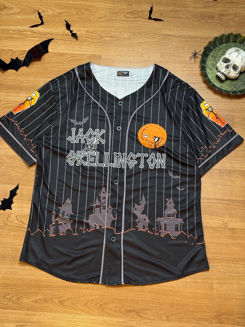 May include: Black baseball jersey with white vertical stripes, featuring the text "Jack Skellington" and a pumpkin design. The jersey has a button-up front and short sleeves. The design includes bats and haunted houses.