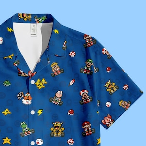 Pode incluir: Camisa azul com botões com um padrão repetido de personagens e elementos pixelados da série de videojogos Mario Kart. O design inclui Mario, Luigi, a Princesa Peach e outros personagens nos seus karts, juntamente com vários power-ups.