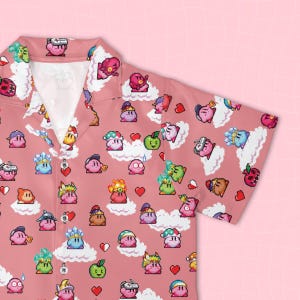 Op de afbeelding: Een overhemd met korte mouwen en knoopsluiting in een oudroze kleur met een all-over pixel art patroon. De print toont verschillende variaties van het personage Kirby, witte wolken, rode harten en groene appels verspreid over de stof.