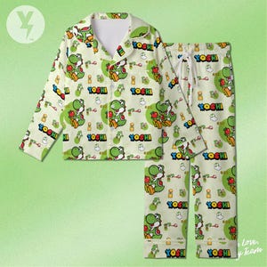 May include: A light cream-coloured pyjama set featuring a repeating pattern of the green character Yoshi, red apples, and the text YOSHI in bold letters. The long-sleeve button-down shirt has a collar and a chest pocket, paired with matching elastic-waist trousers.