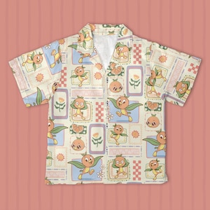 May include: A short-sleeve button-down shirt in a cream color featuring an all-over print of orange bird characters with green wings. The pattern includes rectangular frames, checkered borders, orange slices, and small sun and flower icons.