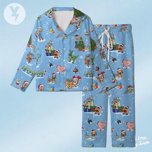 Puede incluir: Un conjunto de pijama de manga larga en color azul claro con un patrón repetitivo de personajes de Toy Story, incluyendo a Woody, Buzz Lightyear, Rex y Slinky Dog con gorros de Papá Noel, junto a árboles de Navidad, regalos y copos de nieve.