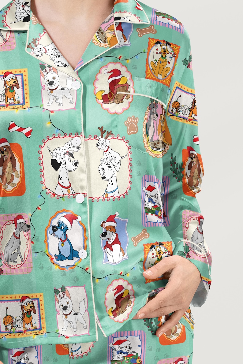 Op de afbeelding: Lichtgroene pyjamatop met een herhalend patroon van cartoonhonden in kerstmutsen en kaders. De pyjamatop heeft witte biezen en knopen. De afbeelding toont een close-up van de pyjamatop.