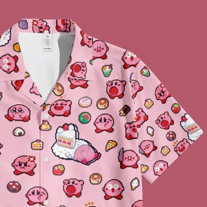Pode incluir: Camisa rosa com botões com um padrão repetido de um personagem de desenho animado rosa e vários alimentos. A camisa de manga curta tem gola de acampamento e interior branco.