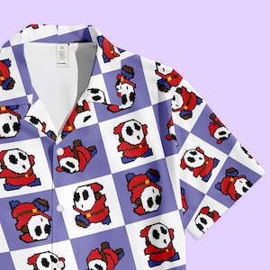 May include: Short-sleeved button-down shirt featuring a repeating pattern of pixelated Shy Guy characters on a checkered background of white and lavender. The shirt has a classic collar and a relaxed fit.
