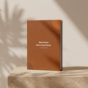 Peut inclure: Un agenda à couverture rigide marron avec le texte "Mental Reset Brain Dump Planner" centré sur la couverture. L'agenda est posé sur une surface claire, avec des ombres de plante en arrière-plan.