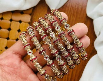 May include: Six gold-toned bangles with red and clear gemstones. The bangles are ornate, with a repeating pattern of red stones and gold embellishments. The jewelry is displayed on a wooden surface with a white cloth in the background.