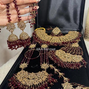 May include: An ornate gold-toned jewelry set with burgundy bead accents, including a necklace, earrings, and bracelet, displayed in a black box. The set features intricate filigree and dangling bead details. The brand name "A & A Zayvraat" is visible.
