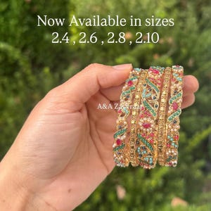 May include: Gold bangles with intricate designs, featuring red, green, and blue gemstones. The text "Now Available in sizes 2.4, 2.6, 2.8, 2.10" is visible. The bangles are held in a hand against a blurred green background.