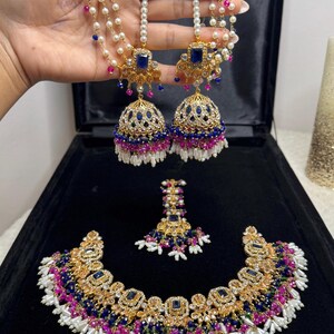 May include: A gold-toned jewelry set featuring a necklace, a pair of jhumka earrings with multi-strand pearl chains, and a maang tikka. The pieces are adorned with rectangular blue stones, small pink and blue beads, and white pearls.