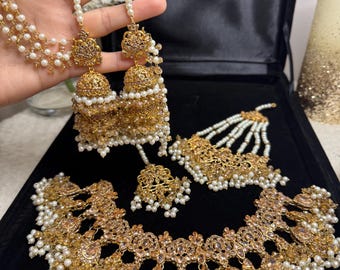 Pode incluir: Um conjunto de joias douradas com colar, brincos e um adorno de cabeça. O conjunto inclui um colar com um design central e elementos pendurados, brincos com um design em forma de sino e um adorno de cabeça com detalhes de pérolas. As joias são adornadas com pequenas pérolas brancas.