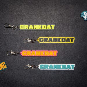 Puede incluir: Cuatro llaveros con la palabra "CRANKDAT" en amarillo, rosa, verde y azul. Cada llavero tiene un cierre plateado. Otras pegatinas con logotipos están dispersas alrededor de los llaveros.