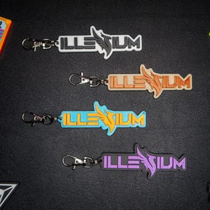 May include: Keychains with the word "ILLENIUM" in white, orange, blue, and purple, each with a matching wing design. The keychains have silver clasps. Other items in the image include a cassette tape and a green word.