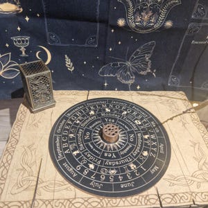 Könnte beinhalten: Ein schwarzes und goldenes Wahrsagebrett mit einem Drehregler mit Buchstaben, Zahlen und Tierkreiszeichen. Ein Metall-Räucherstäbchenhalter und eine dekorative Metallbox sind ebenfalls vorhanden. Der Hintergrund umfasst einen dunklen Stoff mit einem Schmetterlingsdesign.