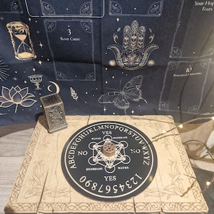 May include: A beige Ouija board with a black planchette and a decorative metal incense burner. The board has the alphabet, numbers, and the words "yes," "no," and "maybe." A dark blue tapestry with gold illustrations is in the background.