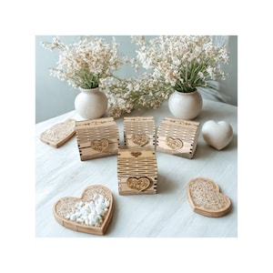 Può includere: Una collezione di scatole di legno e vassoi a forma di cuore. Le scatole hanno un design a righe con un abbellimento a forma di cuore. I vassoi sono riempiti di pietre bianche. Due piccoli vasi bianchi con fiori bianchi sono sullo sfondo.