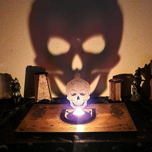 Pode incluir: Um castiçal de madeira em forma de caveira com uma vela acesa, projetando uma grande sombra de caveira na parede. A cena inclui uma tábua Ouija, caixas decorativas e estatuetas, criando um tema de Halloween assustador.