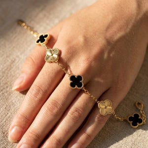 Puede incluir: Pulsera dorada con abalorios en forma de trébol con centros negros y crema. La pulsera está compuesta por una delicada cadena y se muestra en una mano.