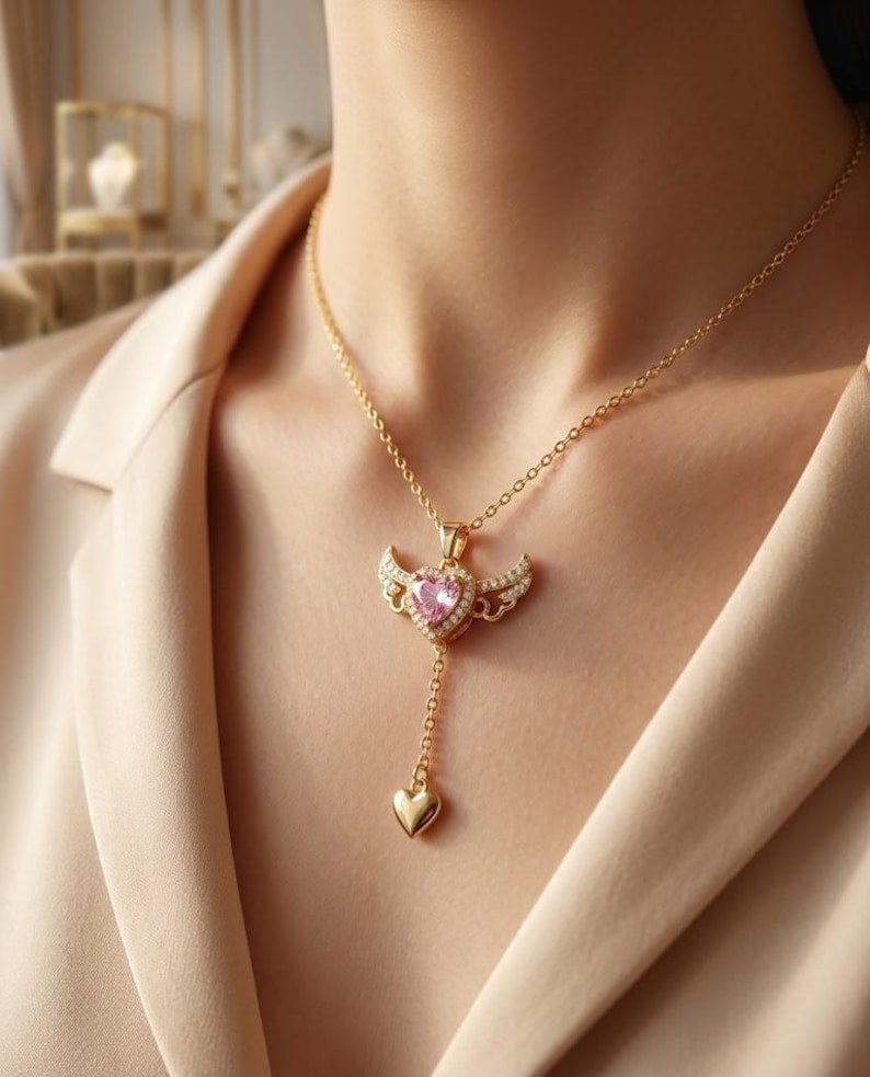 May include: A gold-colored necklace featuring a pendant with a pink heart-shaped gemstone, surrounded by small clear stones and wing-shaped accents. A small gold heart dangles below the pendant. The necklace is worn on a person wearing a beige top.