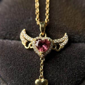 Puede incluir: Un collar dorado con un colgante en forma de coraz&oacute;n. El colgante presenta una piedra preciosa rosa rodeada de peque&ntilde;as piedras transparentes y alas doradas. Un coraz&oacute;n dorado m&aacute;s peque&ntilde;o cuelga debajo, suspendido por una delicada cadena.