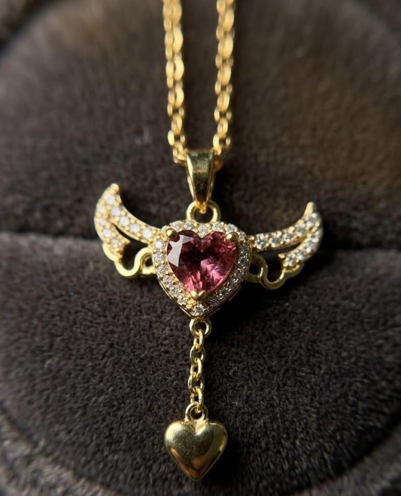 Puede incluir: Un collar dorado con un colgante en forma de coraz&oacute;n. El colgante presenta una piedra preciosa rosa rodeada de peque&ntilde;as piedras transparentes y alas doradas. Un coraz&oacute;n dorado m&aacute;s peque&ntilde;o cuelga debajo, suspendido por una delicada cadena.