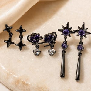 Puede incluir: Un conjunto de tres pares de pendientes de metal negro. Un par presenta estrellas de cuatro puntas, otro par muestra piedras moradas en forma de corazón con anillos orbitales y colgantes claros, y el tercero tiene estrellas con piedras moradas.