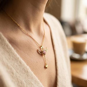 Puede incluir: Un collar dorado con un colgante con una gema rosa en forma de coraz&oacute;n, peque&ntilde;as alas y un coraz&oacute;n colgante. El collar lo lleva una persona que lleva un su&eacute;ter color crema.