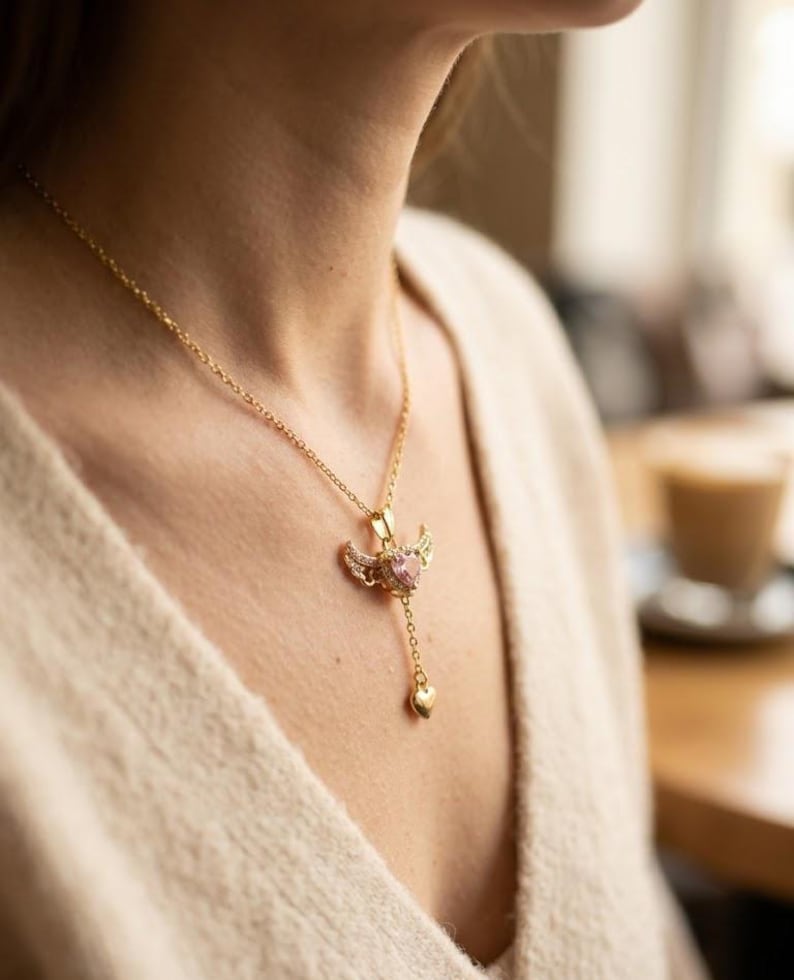 Puede incluir: Un collar dorado con un colgante con una gema rosa en forma de coraz&oacute;n, peque&ntilde;as alas y un coraz&oacute;n colgante. El collar lo lleva una persona que lleva un su&eacute;ter color crema.