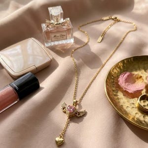 Puede incluir: Un collar dorado con un colgante con una gema rosa en forma de coraz&oacute;n y peque&ntilde;as alas. La imagen tambi&eacute;n muestra un frasco de perfume, maquillaje y un peque&ntilde;o plato dorado con p&eacute;talos de rosa, creando una est&eacute;tica femenina.