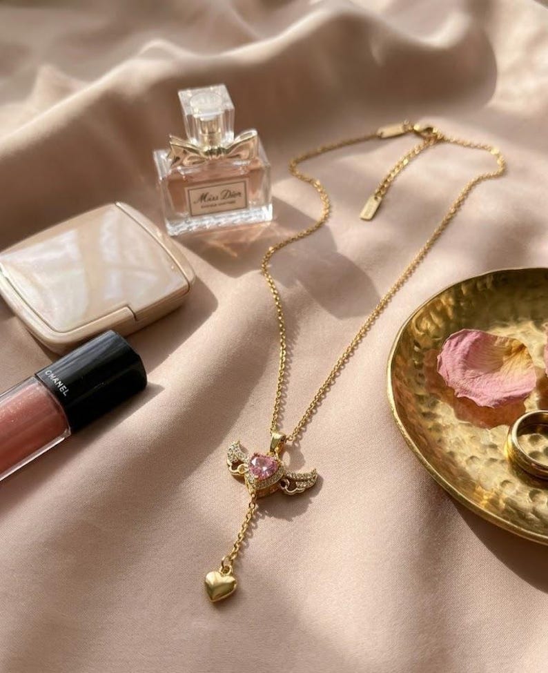 Puede incluir: Un collar dorado con un colgante con una gema rosa en forma de coraz&oacute;n y peque&ntilde;as alas. La imagen tambi&eacute;n muestra un frasco de perfume, maquillaje y un peque&ntilde;o plato dorado con p&eacute;talos de rosa, creando una est&eacute;tica femenina.