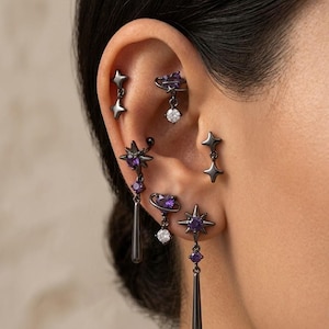 Può includere: Un assortimento di orecchini neri con design a stella e pianeta, adornati con pietre preziose viola e trasparenti. Gli orecchini includono borchie, pendenti e cerchi, creando un tema celeste.