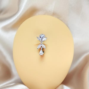 May include: A silver-toned belly button ring with clear, sparkling cubic zirconia stones. The design features a large round stone at the top, with a cluster of smaller stones and a teardrop-shaped stone hanging below. The jewelry is set against a light yellow background.