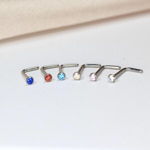 May include: A collection of six L-shaped nose studs with colorful, round gemstones. The studs are silver-toned, and the gemstones are blue, red, light blue, iridescent, pink, and white. The jewelry is displayed on a white surface.