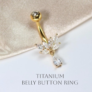 May include: Gold-colored belly button ring with a curved barbell design. The ring features a clear, round gem at the top and a cluster of clear, marquise-cut gems forming a floral shape. A teardrop-shaped gem hangs below. Text reads: TITANIUM BELLY BUTTON RING.