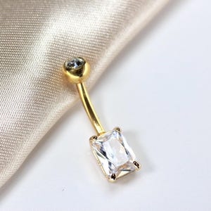 May include: A gold-colored belly button ring with a clear, rectangular-cut gemstone at the bottom and a small, round clear gemstone at the top. The jewelry is set against a soft, cream-colored fabric.