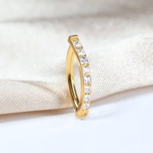 May include: A gold-colored ring with a curved design, set with a row of square, clear gemstones. The ring is displayed against a soft, off-white fabric background, highlighting its sparkle and design.