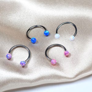 May include: Four horseshoe-shaped body jewelry pieces with opal-like orbs in blue, white, and pink. The jewelry is silver-toned and rests on a satin fabric.