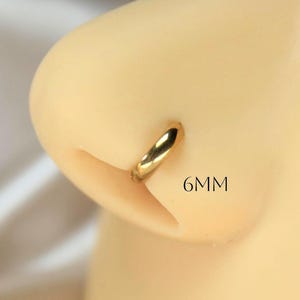 May include: A close-up of a gold-colored nose ring, 6mm in size, worn on a person's nose. The ring is a simple, circular design. The text "6MM" is visible next to the nose ring.