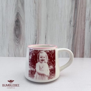 Puede incluir: Una taza de cer&aacute;mica blanca con un borde rosa y una impresi&oacute;n fotogr&aacute;fica monocrom&aacute;tica de color marr&oacute;n rojizo de un ni&ntilde;o sonriente en el frente. La taza tiene un asa curva y un acabado liso, mostrando una imagen clara de una persona.